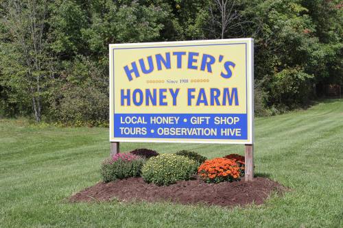 Sign for Hunter's Honey Farm, featuring local honey, a gift shop, tours, and an observation hive, surrounded by flowers.