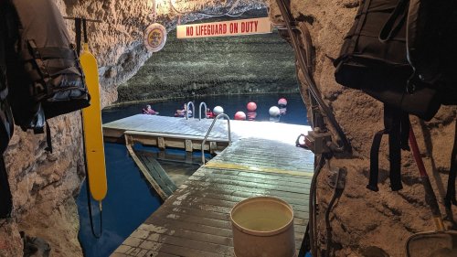 View of a cave entrance with a wooden dock, swimming area, and a "No Lifeguard on Duty" sign above.