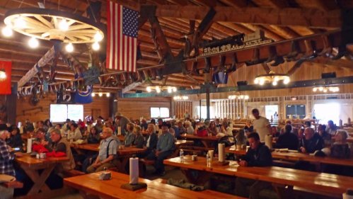 A large group of people seated at wooden tables in a rustic hall, with an American flag and wooden beams overhead.
