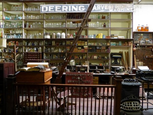 A vintage store interior with shelves of jars, tools, and a wooden ladder, showcasing historical items and machinery.