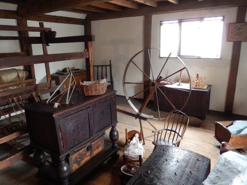 A rustic room with wooden furniture, a spinning wheel, and various vintage items, illuminated by natural light.