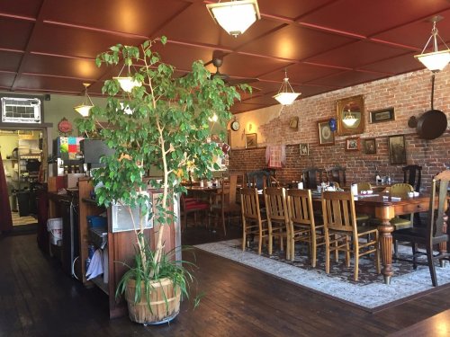 Cozy restaurant interior with wooden tables, chairs, a potted plant, and brick walls adorned with framed pictures.