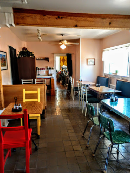Cozy restaurant interior with wooden tables, colorful chairs, and a warm atmosphere. Brightly lit with a homey feel.