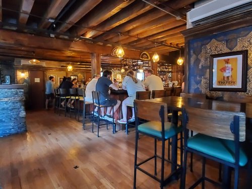A cozy restaurant interior with wooden beams, patrons seated at the bar, and warm lighting creating a welcoming atmosphere.