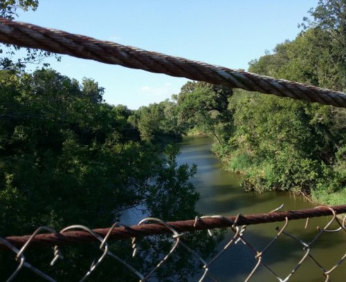 A serene river flows through lush greenery, viewed from a bridge with rusted cables and a chain-link fence.