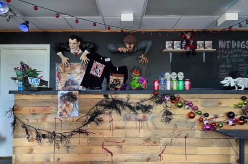 A spooky-themed counter decorated with Halloween props, shirts, and colorful drinks, set in a dimly lit space.