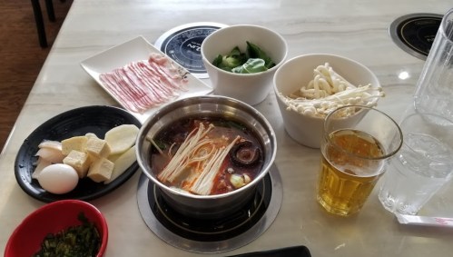 A table set with a hot pot, various ingredients like vegetables, meat, and eggs, and a glass of beer.