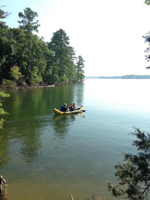 Two people kayak on a calm lake surrounded by lush green trees under a clear blue sky.