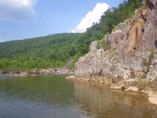 A serene river scene with rocky cliffs and lush green hills under a blue sky with scattered clouds.