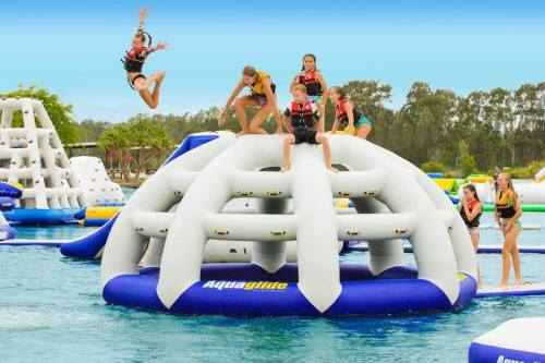 Children in life jackets play on an inflatable water structure, with one jumping off and others climbing on it.