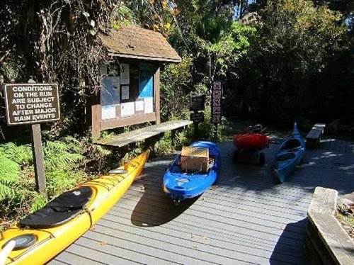 A wooden dock with kayaks in orange and blue, surrounded by lush greenery and a sign about water conditions.