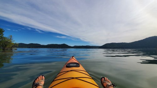 A view from a kayak on calm water, with mountains and a blue sky in the background. Feet resting on the kayak's edge.
