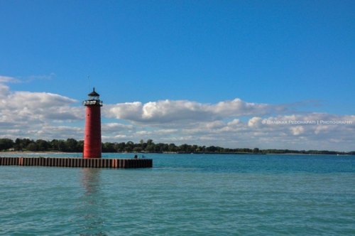 A red lighthouse stands on a pier by calm blue waters under a partly cloudy sky.