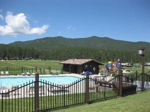 A sunny pool area with a wooden building, surrounded by a fence and mountains in the background.