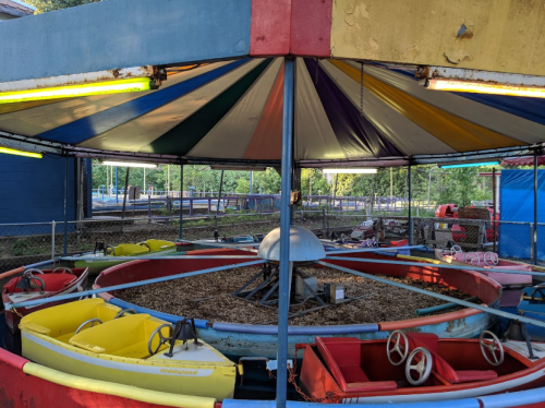 A colorful carousel with empty boats under a striped canopy, surrounded by a fence in an amusement park setting.
