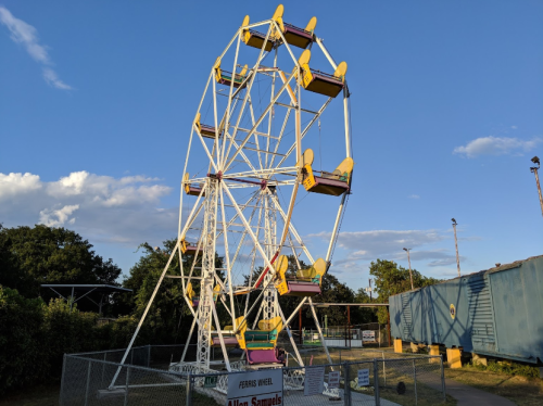 A colorful Ferris wheel stands against a blue sky, surrounded by a fence and a shipping container nearby.