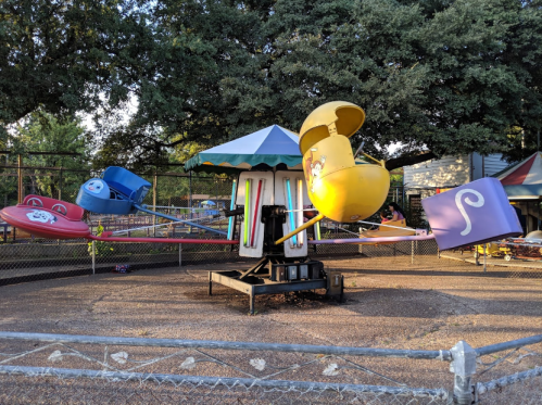 A colorful amusement ride with spinning cars under a shaded canopy, surrounded by a fence and trees in the background.