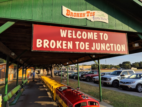 A colorful sign reads "Welcome to Broken Toe Junction" above a small train at a station with parked cars in the background.