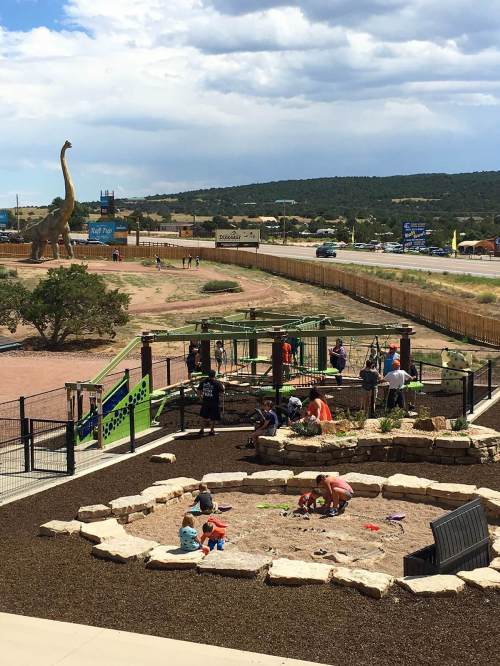 Children play in a sandy area at a park, with a dinosaur statue in the background and a playground nearby.