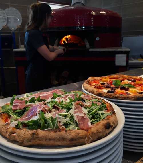 A stack of pizzas topped with arugula and ham, with a pizza oven and a person in the background preparing food.