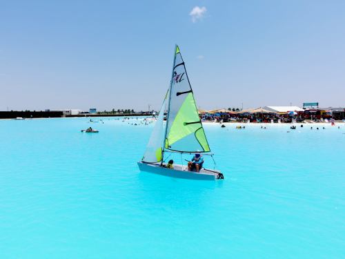 A small sailboat with two people glides across a bright turquoise lagoon, surrounded by beachgoers enjoying the sun.