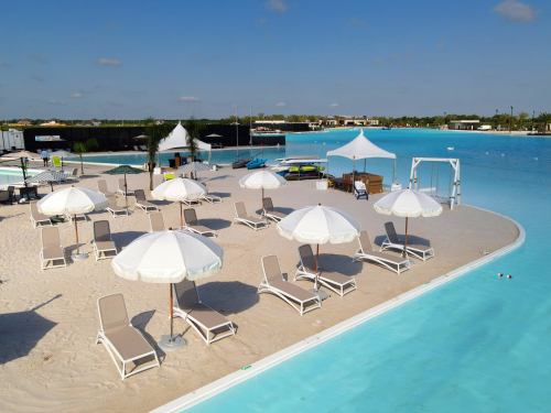 A sunny beach area with white umbrellas, lounge chairs, and a clear blue pool in the background.