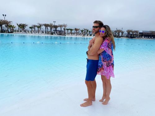 A couple stands together by a large pool, smiling and enjoying the moment, with palm trees in the background.