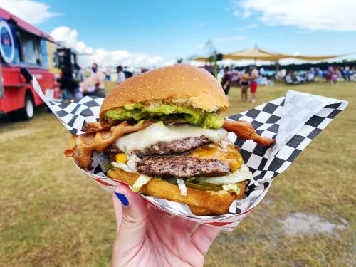 A hand holding a large burger with bacon, cheese, and toppings, set against a food truck and outdoor event backdrop.