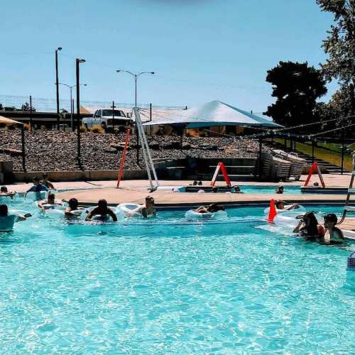 A sunny pool scene with people relaxing on floats and lounging by the water, surrounded by trees and a clear blue sky.