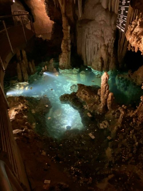 A dimly lit cave with a turquoise pool, surrounded by stalagmites and rock formations.