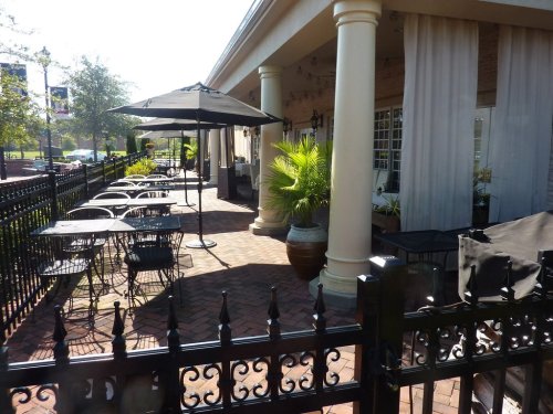 Outdoor patio with black metal tables and chairs, umbrellas, and potted plants, surrounded by a decorative fence.