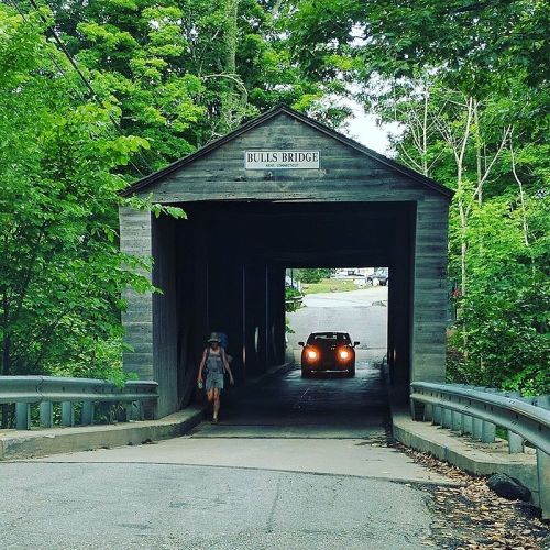 A car drives through a wooden covered bridge while a person walks nearby, surrounded by lush green trees.