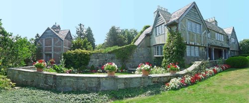 A picturesque stone house surrounded by lush greenery and colorful flower beds under a clear blue sky.
