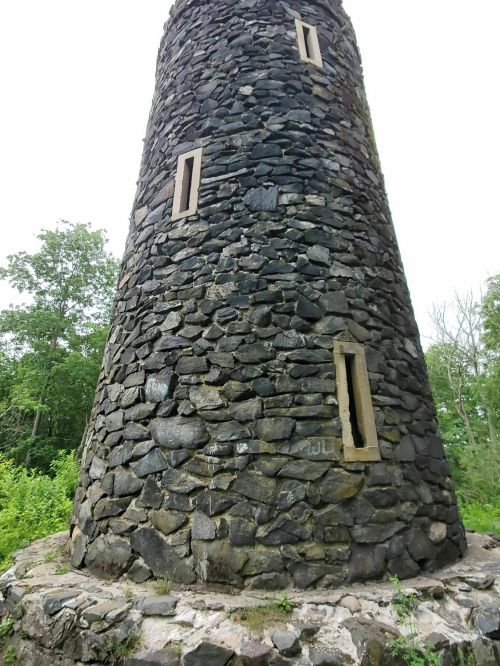 A tall stone tower with narrow windows, surrounded by greenery and trees under a cloudy sky.