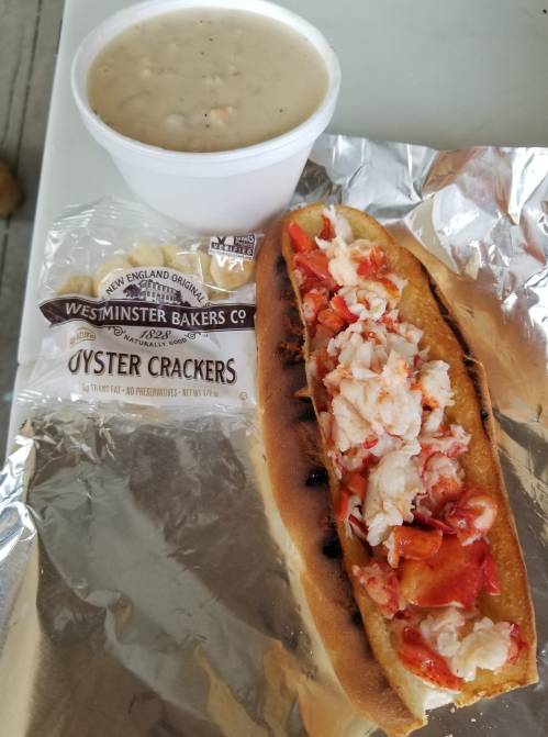 A lobster roll on a foil wrapper next to a cup of clam chowder and a pack of oyster crackers.