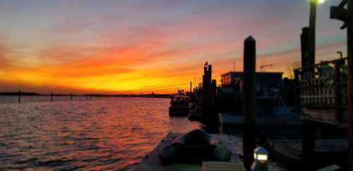 A serene sunset over a calm waterway, with boats docked and silhouettes of posts against a colorful sky.