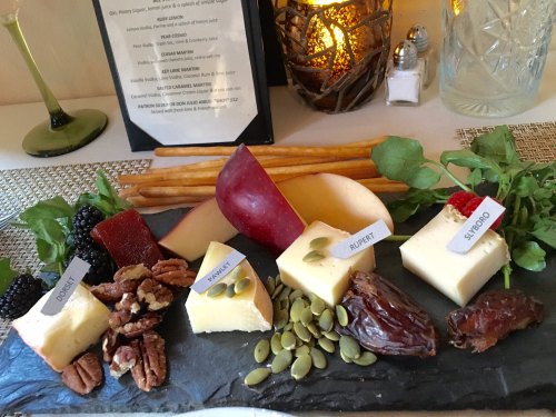 A cheese platter featuring various cheeses, fruits, nuts, and seeds, arranged on a slate board with labels.