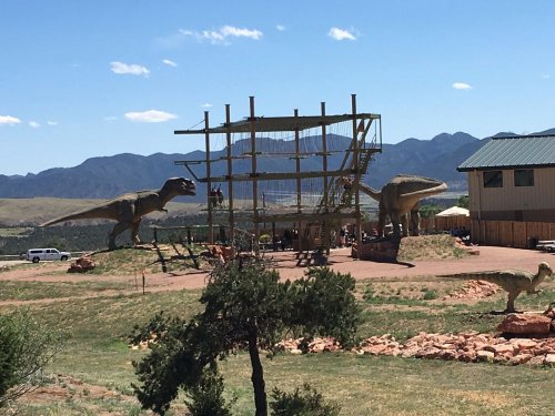 A dinosaur-themed park featuring large dinosaur sculptures and a climbing structure against a mountain backdrop.