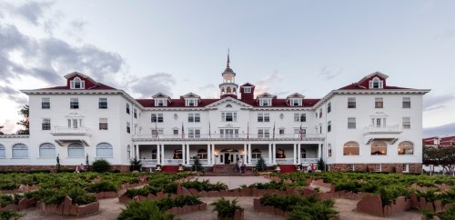 Historic white hotel with a red roof, surrounded by greenery and a cloudy sky.