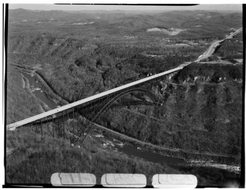Aerial view of a large bridge spanning a river, surrounded by mountains and dense forest. Black and white image.