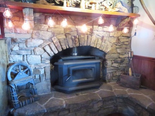 A rustic stone fireplace with a wood stove, surrounded by warm string lights and wooden decor.