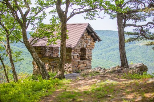 A stone cabin with a metal roof surrounded by trees, overlooking a lush green mountain landscape.