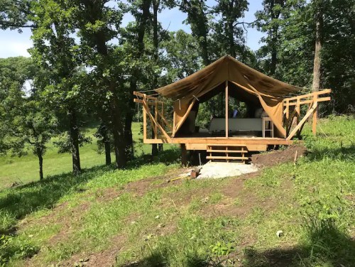 A wooden tent structure on a grassy hillside, surrounded by trees and open fields under a clear blue sky.