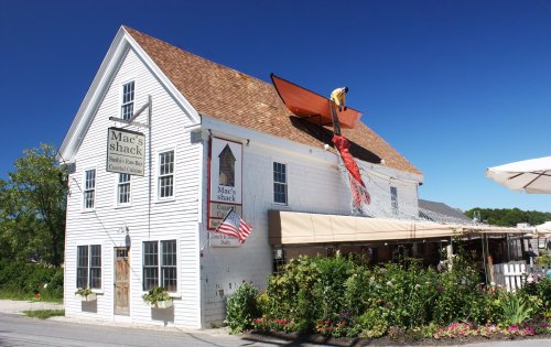 A white building with a sign reading "Mac's Shack," featuring a person working on the roof under a clear blue sky.