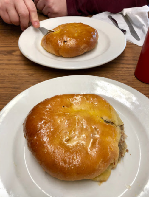Two golden, cheesy meat pies on white plates, one being cut with a fork, on a wooden table.