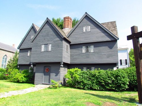 A large, gray, historic house with a unique roof design, surrounded by greenery and a clear blue sky.