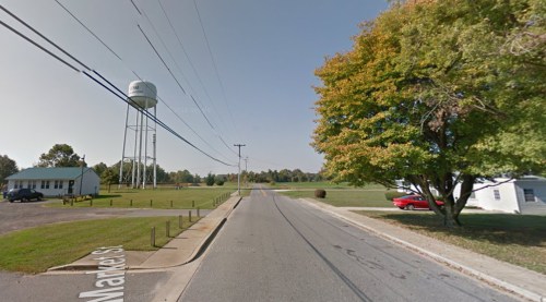 A quiet street scene featuring a water tower, trees, and a red car parked near a grassy area.