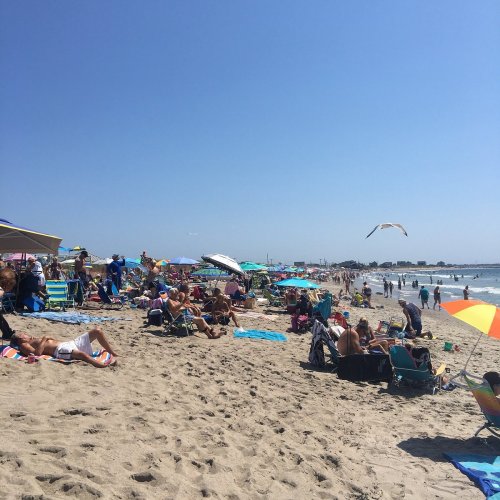 Crowded beach scene with sunbathers, umbrellas, and colorful beach chairs under a clear blue sky.