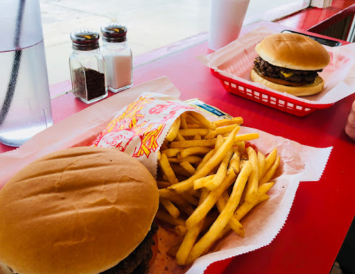 Two burgers on red trays with a side of golden fries, set on a red table with condiments in the background.