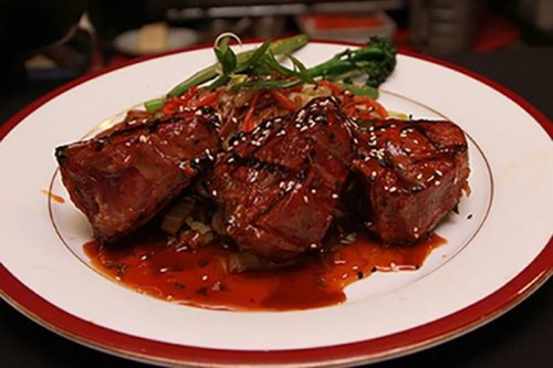 Grilled steak pieces on a plate, garnished with vegetables and a drizzle of sauce, topped with sesame seeds.